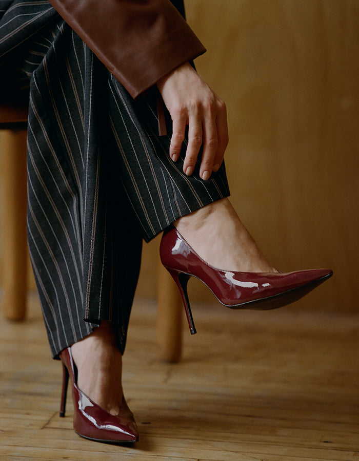 A person seated indoors wearing glossy burgundy pointed-toe high heels with slim stiletto heels and dark pinstriped trousers, against a warm wood background.