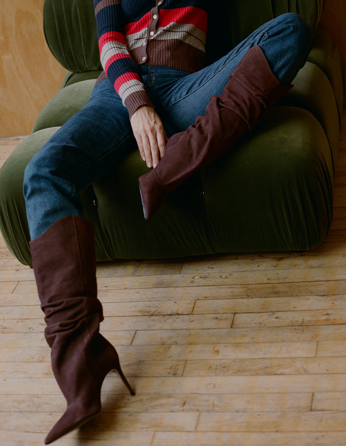 Person seated on a dark green velvet armchair wearing blue jeans, a striped cardigan, and brown suede slouchy high-heeled knee-high boots