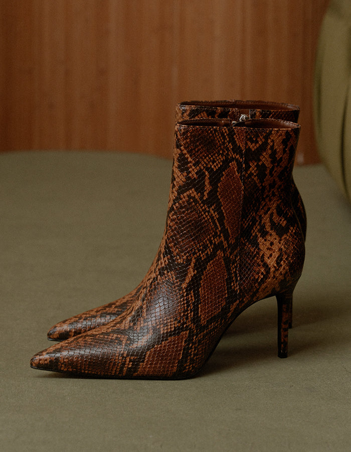 A pair of pointed-toe ankle boots with a brown and black snakeskin pattern, featuring a mid-height stiletto heel, displayed on a neutral surface against a wooden background.