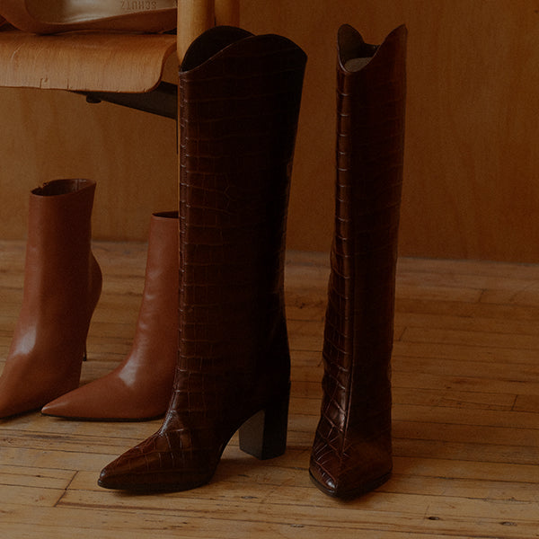 Pair of tall dark brown croc-embossed leather boots standing on a wooden floor, with tan leather ankle boots in the background.