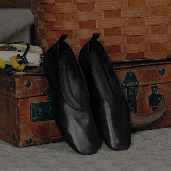 Pair of black leather ballet flats leaning against a vintage suitcase and a woven basket.