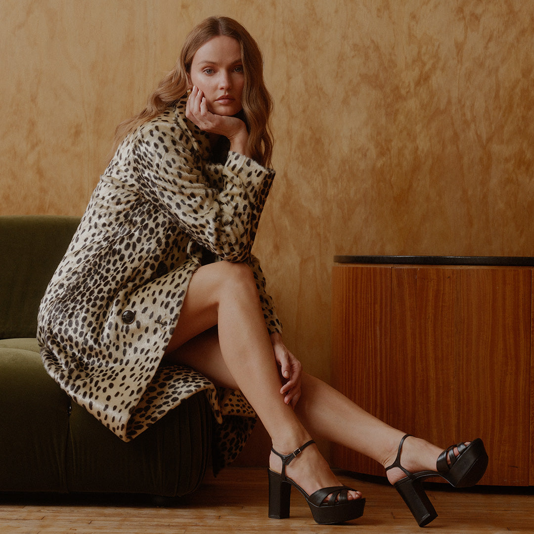 A woman sits on a dark green velvet sofa wearing a beige and black leopard-print coat and black platform sandals, posed in front of a textured warm brown wall.