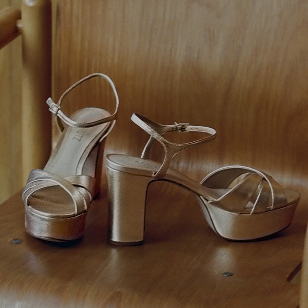 Pair of metallic champagne-gold platform sandals with block heels on a wooden chair.