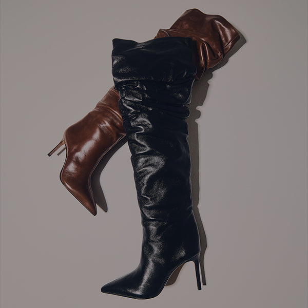 Two tall boots arranged side by side against a neutral wall. One boot is crafted in rich brown leather, and the other in glossy black leather. Both have pointed toes and high heels, shown under angled lighting that emphasizes their texture.