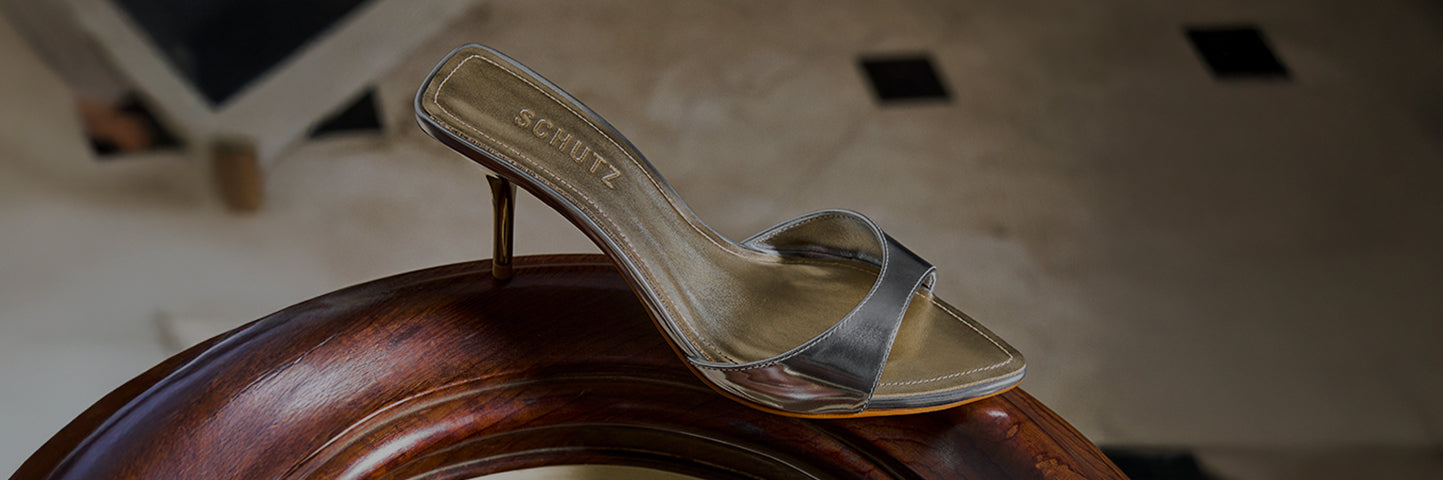 A metallic heeled mule with a slim strap resting on a polished wooden surface, featuring the SCHUTZ logo on the insole.