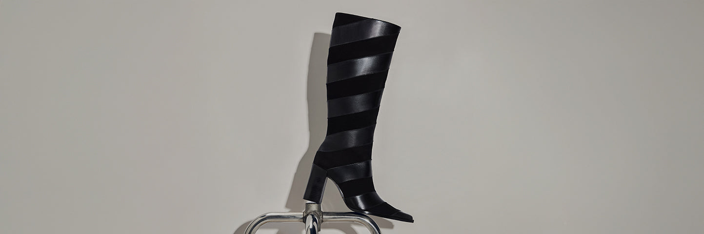 Black knee-high boots with alternating matte and glossy leather stripes, displayed on a chair.