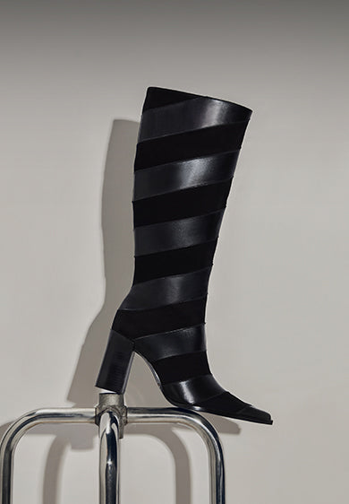 Black knee-high boots with alternating matte and glossy leather stripes, displayed on a chair.