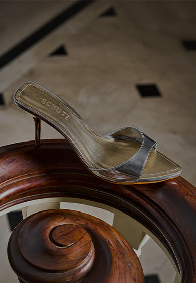 A metallic heeled mule with a slim strap resting on a polished wooden surface, featuring the SCHUTZ logo on the insole.