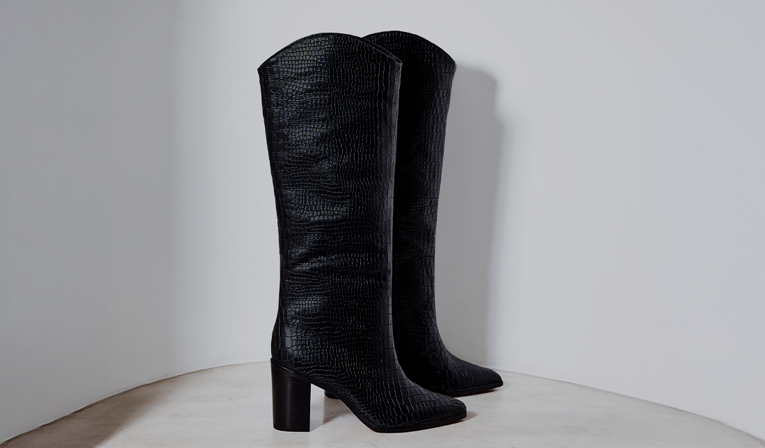 Essentials – Tall black boots with a textured, slightly glossy finish standing upright against a clean white wall. The design is structured and elegant, giving a timeless, essential wardrobe feel.