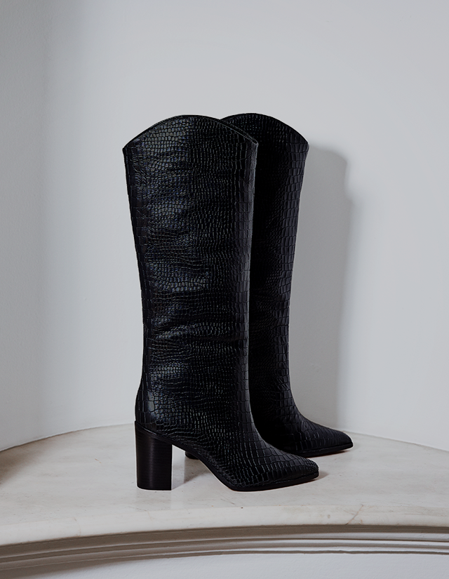 Essentials – Tall black boots with a textured, slightly glossy finish standing upright against a clean white wall. The design is structured and elegant, giving a timeless, essential wardrobe feel.