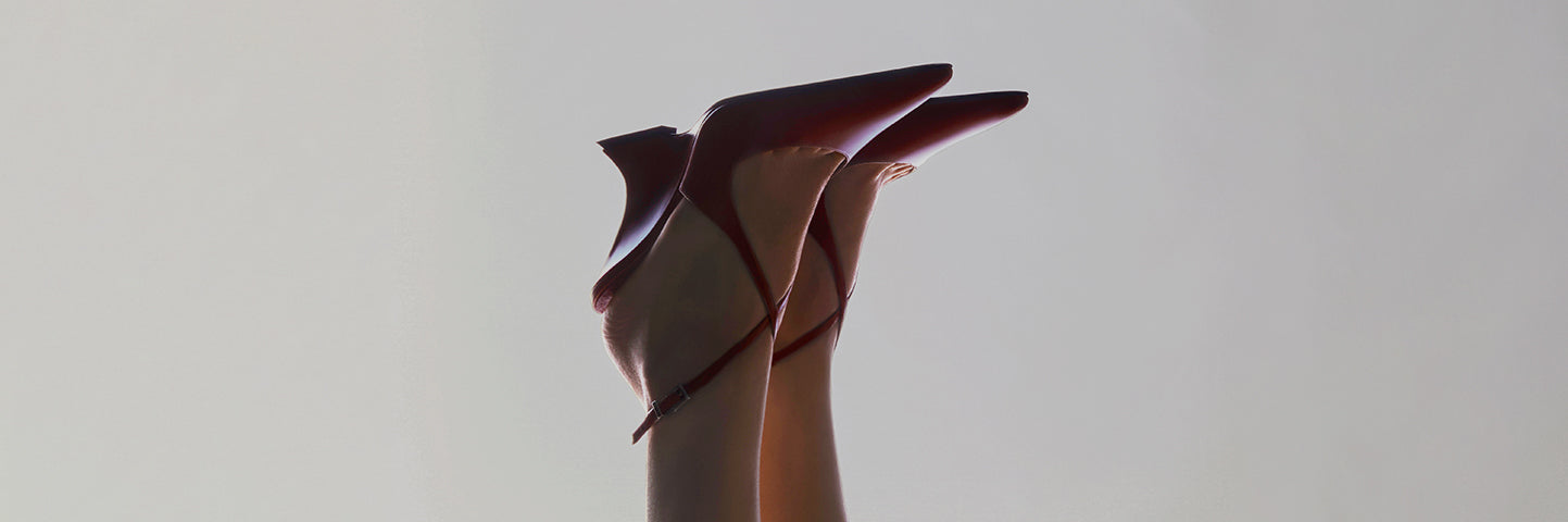 Pair of pointed-toe red heels with ankle straps, lifted in the air against a light background.