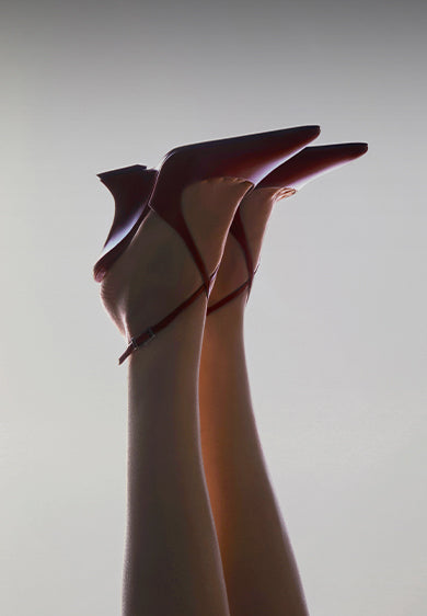 Pair of pointed-toe red heels with ankle straps, lifted in the air against a light background.