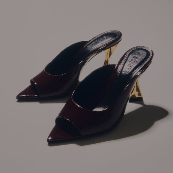 Close-up of a single black patent leather slide mule with a high, thin stiletto heel featuring a gold tone core, viewed from a high angle.
