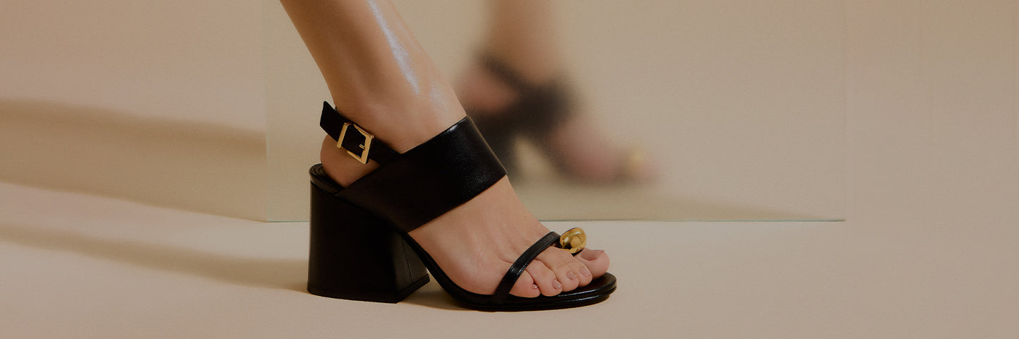 Women’s black leather sandals with a block heel, ankle strap, and subtle gold detail, shown worn on foot in a clean studio environment.