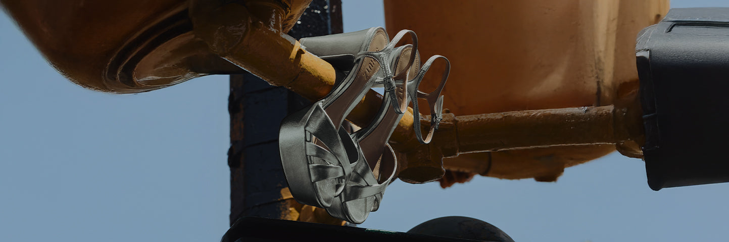 A pair of silver metallic platform sandals with peep toes and high heels, featuring crisscross straps and ankle support, hanging from the yellow bars of an industrial metal structure under a blue sky. The text 'KEEFA SANDAL' is shown on the left.