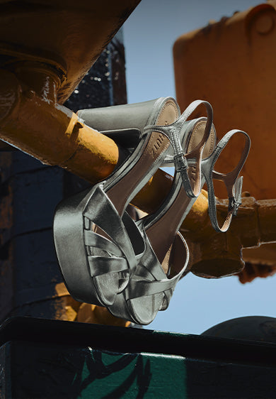 A pair of silver metallic platform sandals with peep toes and high heels, featuring crisscross straps and ankle support, hanging from the yellow bars of an industrial metal structure under a blue sky. The text 'KEEFA SANDAL' is shown on the left.