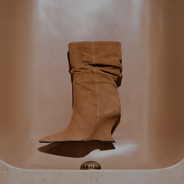 Pair of suede slouchy booties in a warm tan shade, displayed upright on the smooth edge of a bathtub. The boots have a pointed toe and a slim heel, styled in soft natural lighting.
