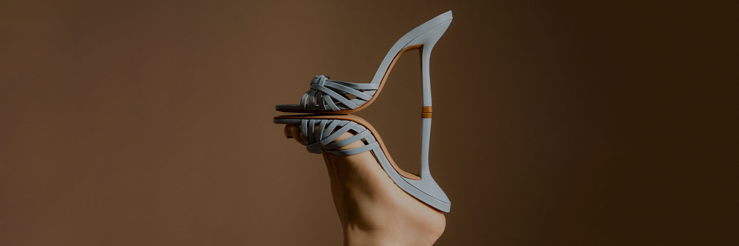 Promotional banner featuring a light blue stiletto sandal with slim straps and a high heel, shown in close-up against a warm gradient background, with the text ‘Stiletto Sandals’ and a subtitle about versatile day-to-night styling.