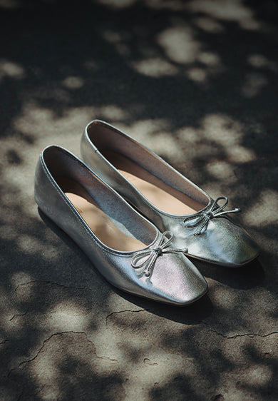 A pair of shiny silver ballet flats with rounded square toes and small decorative bows on top, resting on a textured stone surface dappled with soft shadows from tree leaves. The label 'ARISSA STYLE' is written to the left.
