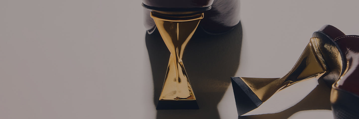 A close-up of the heel area of a pair of dark brown or burgundy shoes with a mid-height, sculptural gold heel. The heel has a unique, hourglass or criss-cross shape, and the shoes cast long shadows on a neutral background. The text MID HEEL STYLES is written in large white letters on the left side of the image."