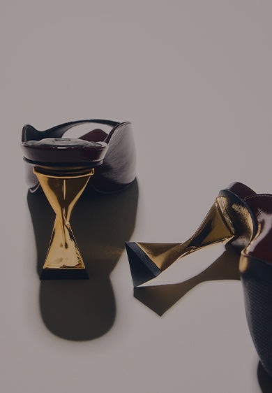 A close-up of the heel area of a pair of dark brown or burgundy shoes with a mid-height, sculptural gold heel. The heel has a unique, hourglass or criss-cross shape, and the shoes cast long shadows on a neutral background. The text MID HEEL STYLES is written in large white letters on the left side of the image."