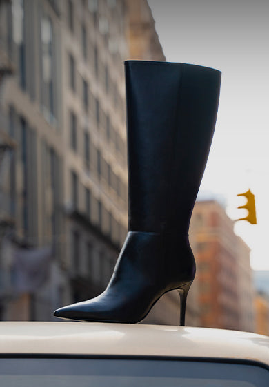 Sleek black Mikki boot with pointed toe and stiletto heel, showcased outdoors on a city street with blurred buildings in the background.