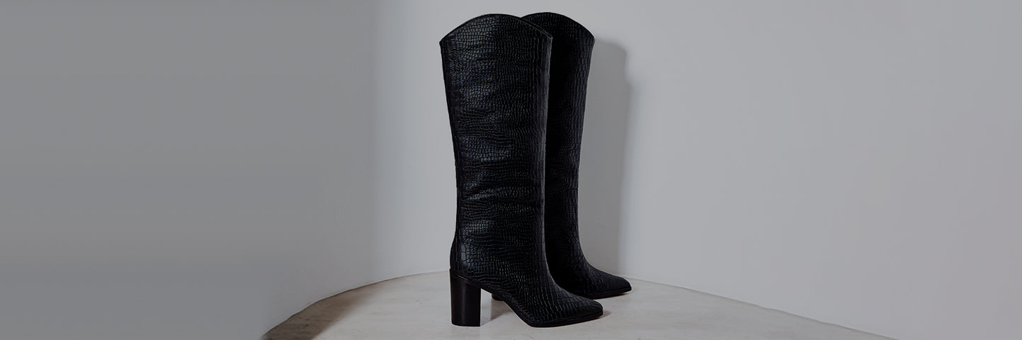 A pair of tall black textured Maryana boots standing upright against a light gray wall.