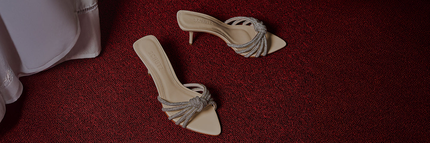 Pair of beige mule sandals with thin high heels and braided metallic straps, placed on a red textured carpet beside a light-colored fabric.