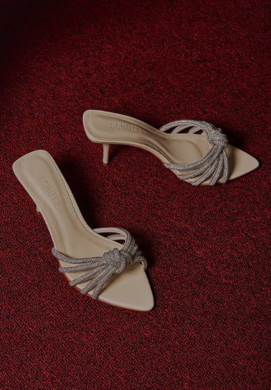 Pair of beige mule sandals with thin high heels and braided metallic straps, placed on a red textured carpet beside a light-colored fabric.
