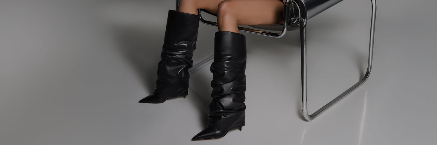 Women wearing black high-heeled boots with pointed toes, seated in a modern interior setting with a monochromatic color palette.