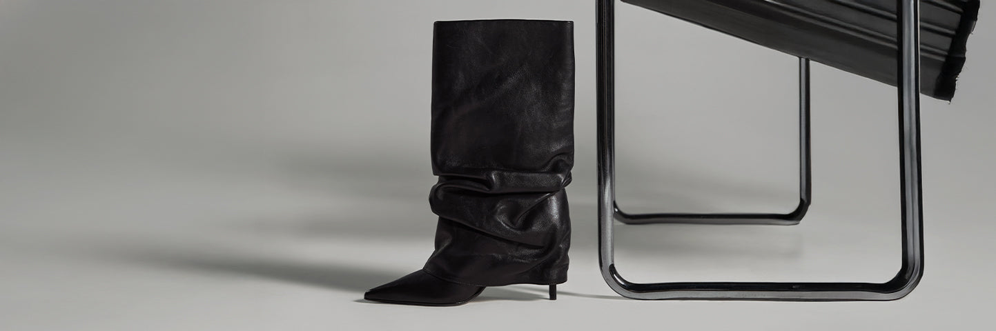 Tall black leather boot with a slouchy design, pointed toe, and slim stiletto heel, photographed against a minimalist modern chair in a studio setting.