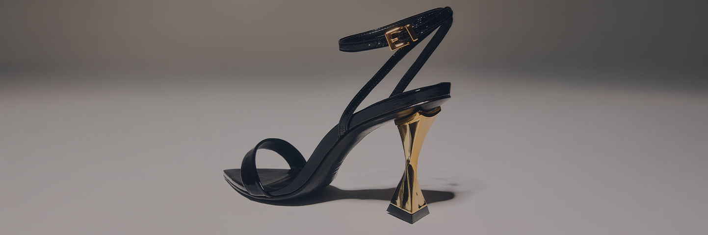Black mid-heel sandal with a sculptural gold geometric heel, displayed on a neutral background with the text ‘MID HEEL SANDALS.’