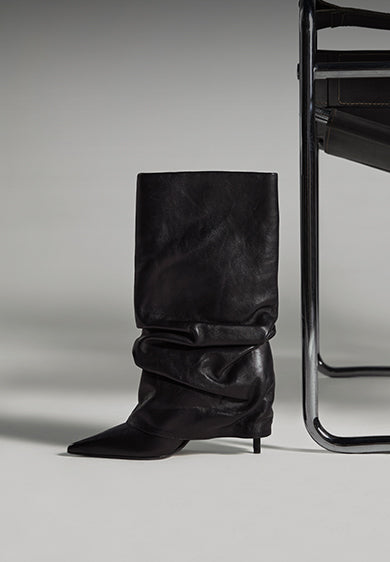 Tall black leather boot with a slouchy design, pointed toe, and slim stiletto heel, photographed against a minimalist modern chair in a studio setting.