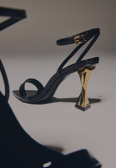 Black mid-heel sandal with a sculptural gold geometric heel, displayed on a neutral background with the text ‘MID HEEL SANDALS.’