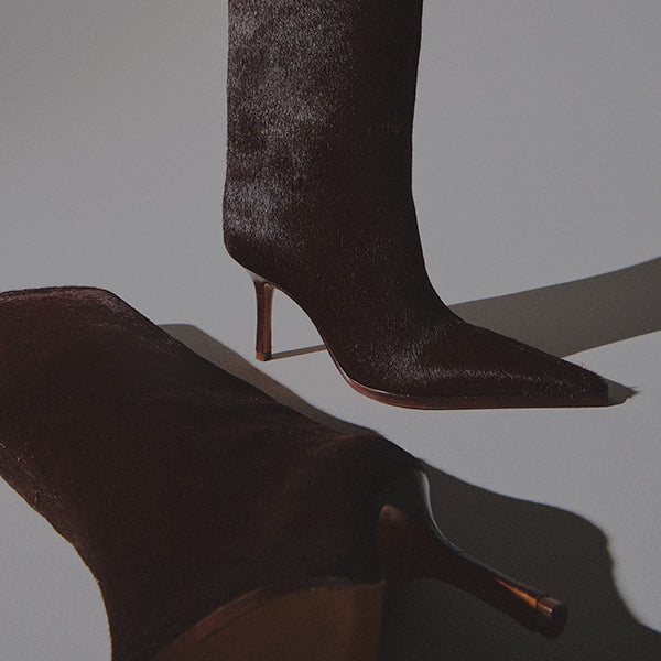 A single dark brown, knee-high boot in textured hair calf material, with a pointed toe and a thick block heel, standing on a light gray table.