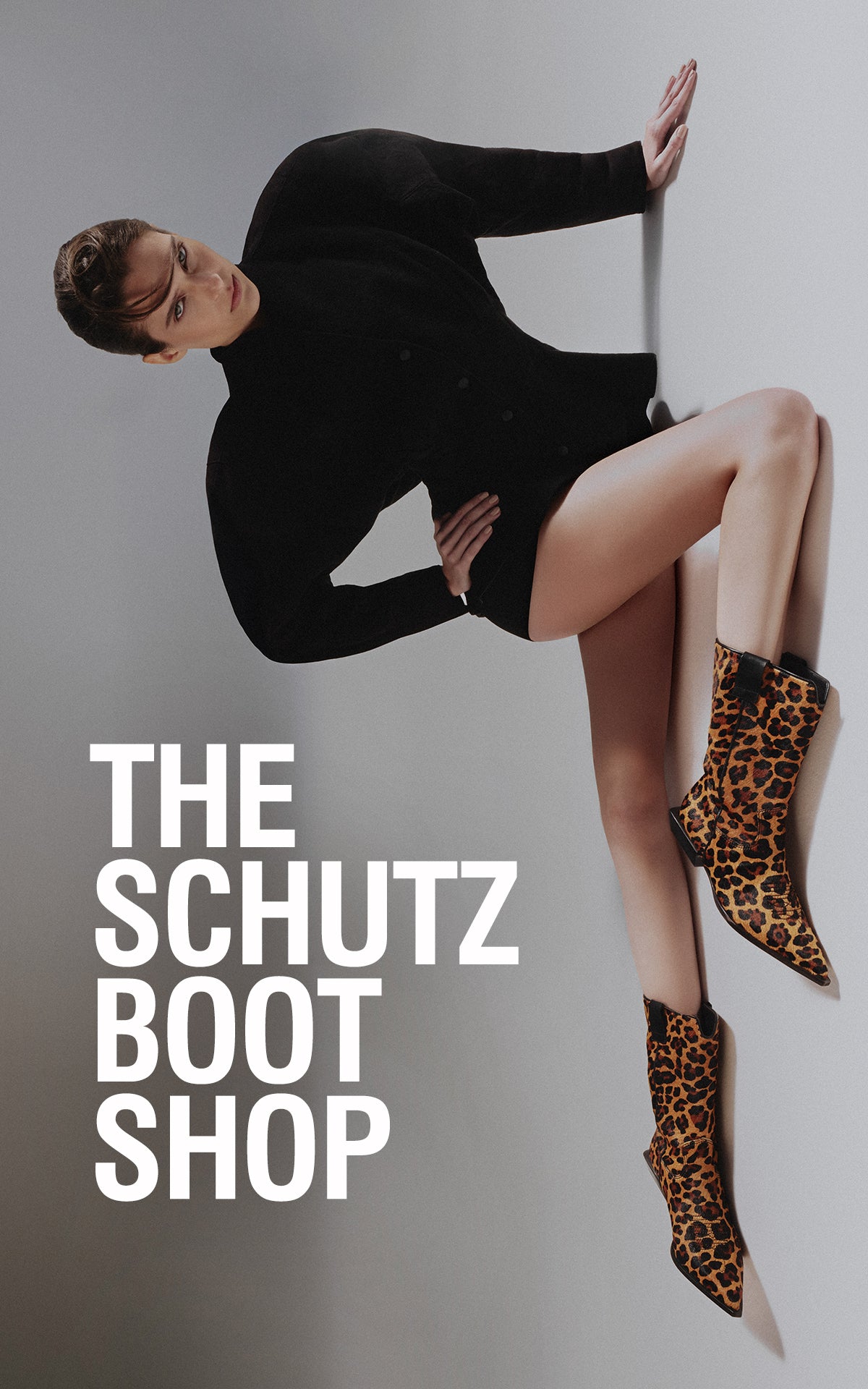 A stylish woman with long, wavy blonde hair poses confidently in an outdoor industrial setting. She wears an oversized black blazer and high-heeled platform sandals, embodying modern elegance. On the right side of the image, a close-up of a woman's legs showcases sleek black knee-high boots as she steps on a metallic ladder, with a background filled with fabric rolls. Bold white text welcomes visitors to Schutz, emphasizing the brand’s reputation for top-tier footwear.