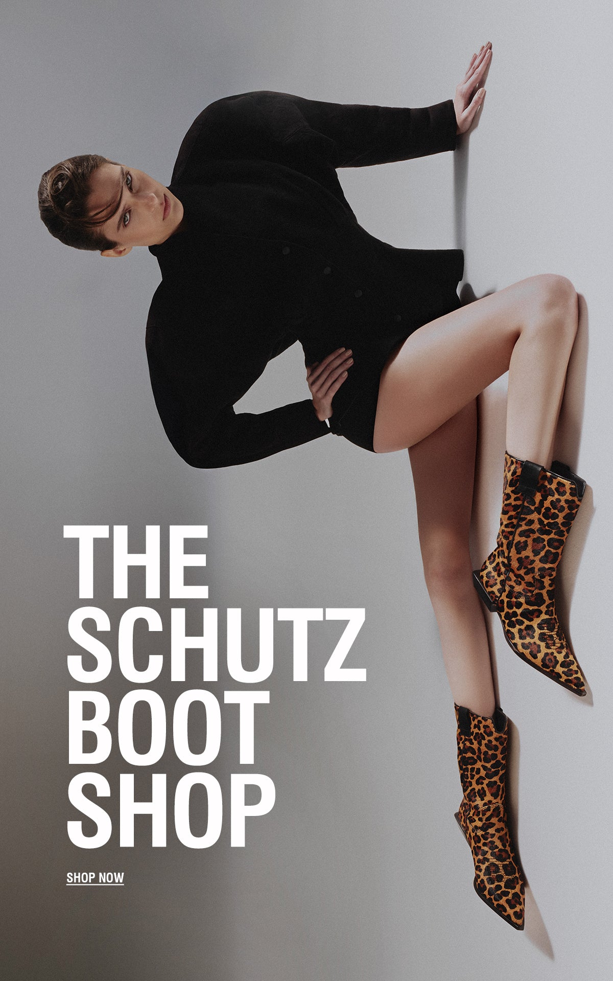 Model wearing a structured black outfit and leopard-print ankle boots, posed on the floor against a minimalist gray background, with large white text reading “THE SCHUTZ BOOT SHOP” and a “SHOP NOW” button.