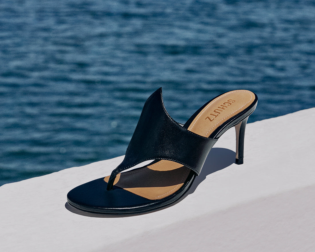 A sleek black heeled mule sandal from Schutz, placed on a white ledge with a blue ocean in the background.