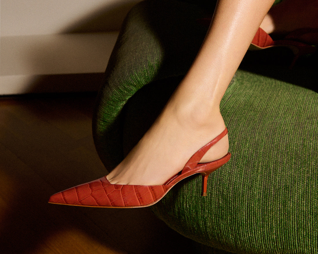 A close-up of a model’s foot in a red, textured slingback pump with a kitten heel. The text reads: "On Trend Pumps – Shop Now."