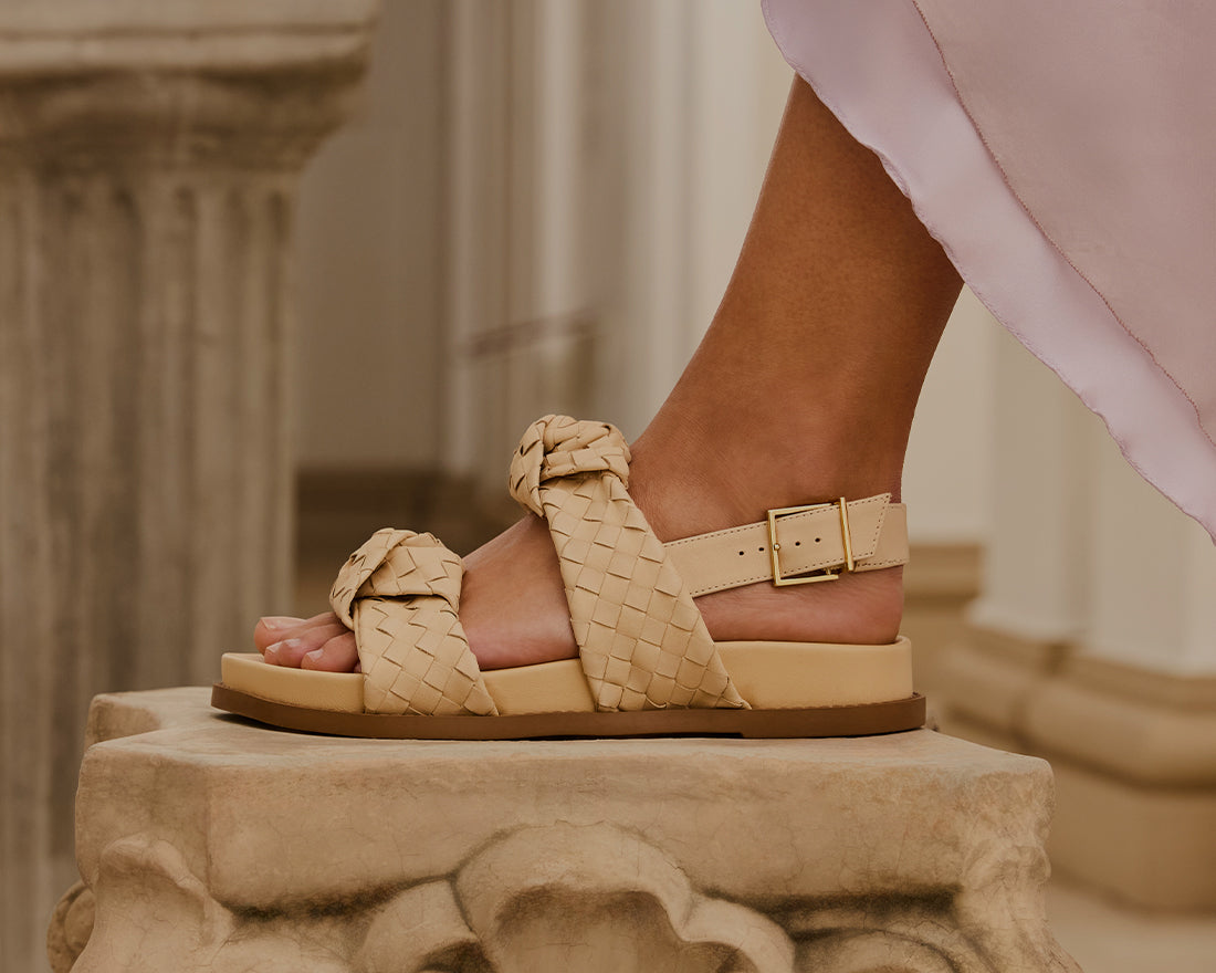 Close-up shot of a model’s foot wearing intricately detailed flat sandals with braided nude straps and a gold buckle, standing next to classic architectural columns.
