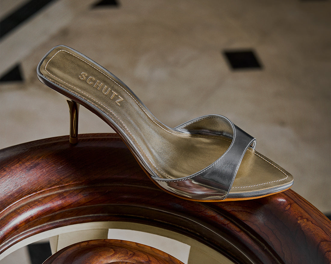 Metallic gold mule sandal with a thin heel resting on a polished wooden surface, with the text ‘Holiday-Ready Sandals.