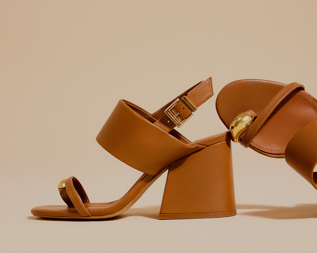 Brown leather heeled sandal with ankle strap and gold detail against a neutral background, with the text “Elevated Versatility” and “Shop Sandals.”