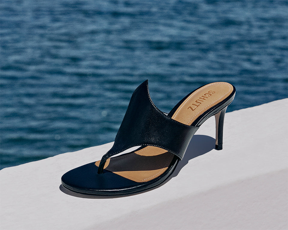 A sleek black heeled mule sandal from Schutz, placed on a white ledge with a blue ocean in the background.