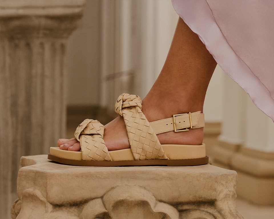 Close-up shot of a model’s foot wearing intricately detailed flat sandals with braided nude straps and a gold buckle, standing next to classic architectural columns.