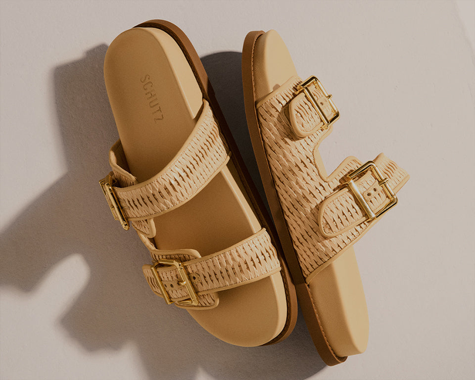 Close-up shot of a model’s foot wearing intricately detailed flat sandals with braided nude straps and a gold buckle, standing next to classic architectural columns.