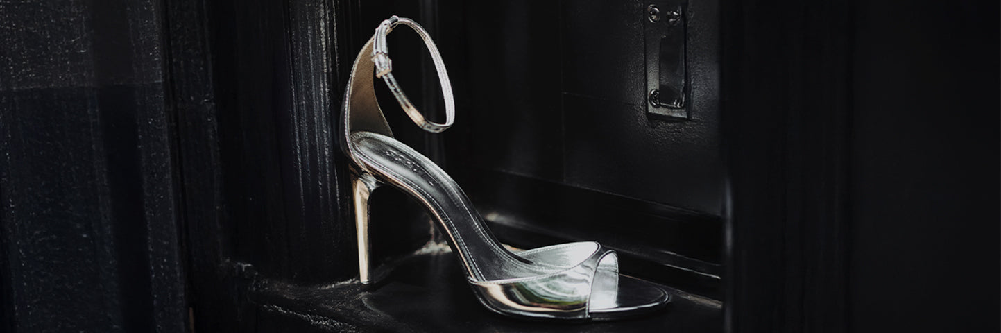 A shiny metallic silver high-heeled sandal with a delicate ankle strap is placed elegantly against a glossy black surface, reflecting light dramatically. Part of the "Metallics" selection.