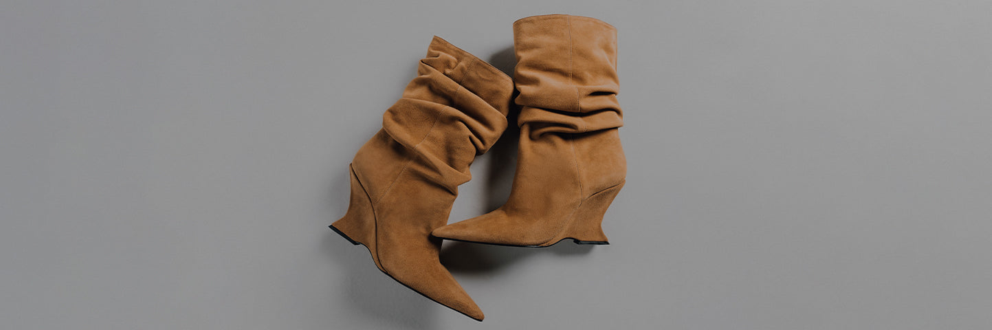 An overhead shot of a pair of slouchy, pointed-toe, tan suede ankle boots (booties) featuring a curved or sculptural wedge heel. The text "BOOTS & BOOTIES" is overlaid on the left side of the gray background.