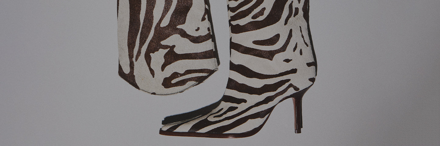 A close-up shot of the lower portion of a pair of high-heeled boots and possibly a matching bag, all made of white and dark brown zebra-print hair-on hide material. The boots have a square toe and a tall, brown stiletto heel. The text "WILD STYLES" is overlaid in the center of the image.