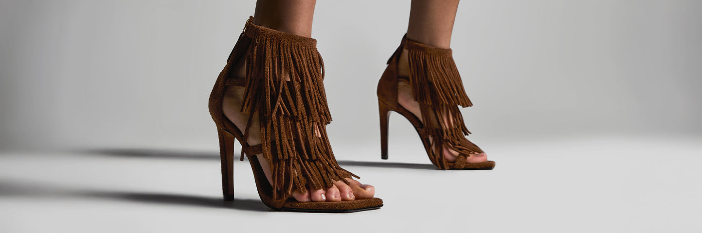 Close-up shot of a woman’s legs wearing high-heeled brown suede sandals adorned with long fringes, evoking a western-inspired style. The shoes stand out against a minimalist gray background, with the bold white text 'WESTERN ROOTS' displayed beside them, emphasizing the collection’s rustic and fashion-forward aesthetic.