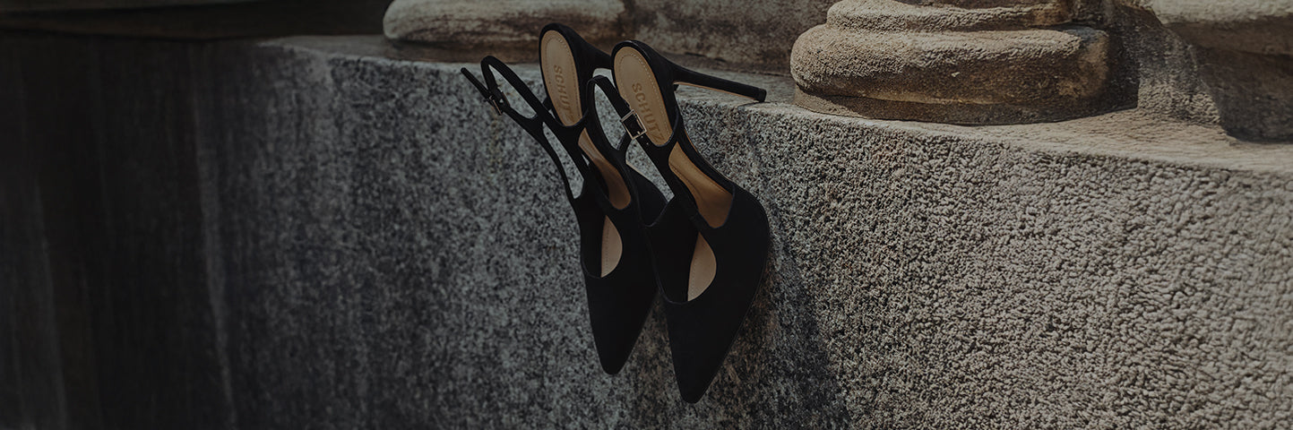 Black pointed-toe slingback heels resting on a stone ledge, with thin straps and a minimalist, elegant design.
